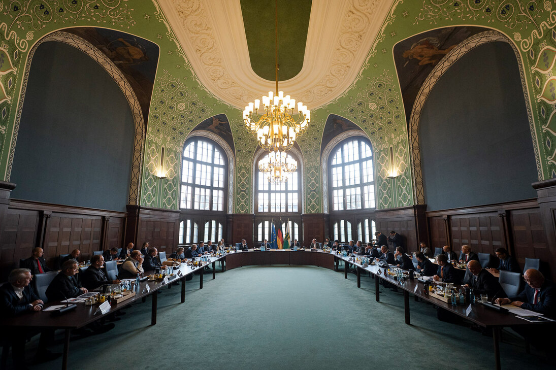 In einem großen Saal sitzen Männer und Frauen an einem Konferenztisch.