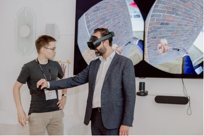 Eine Person trägt eine VR-Brille und links davon steht eine weitere Person und beobachtet das Geschehen