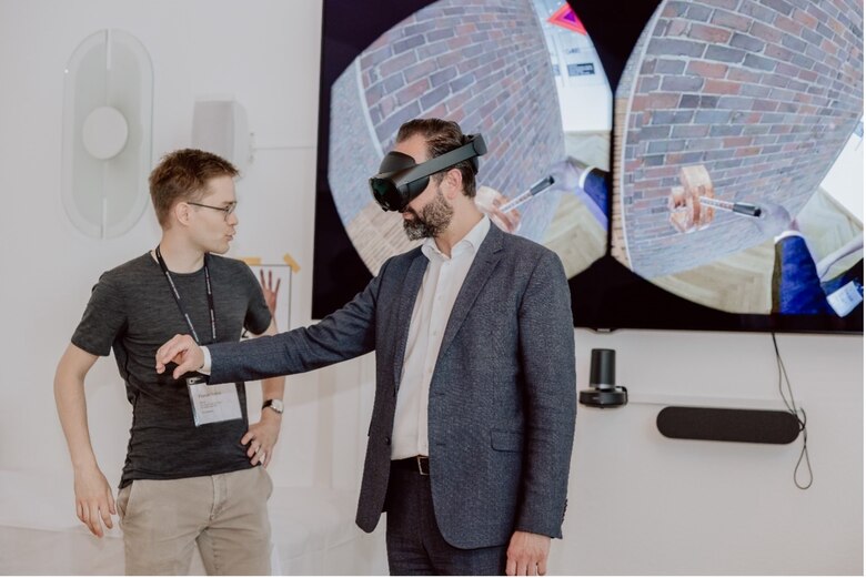 Eine Person trägt eine VR-Brille und links davon steht eine weitere Person und beobachtet das Geschehen