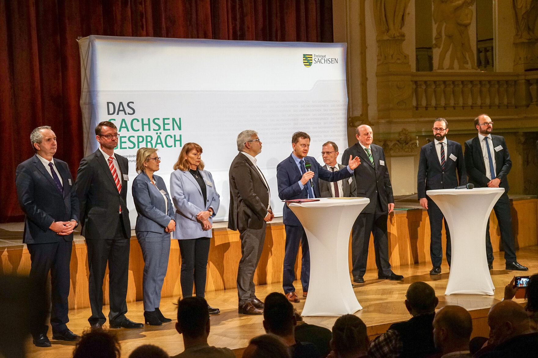 Männer und Frauen stehen in einem Saal auf einer Bühne.