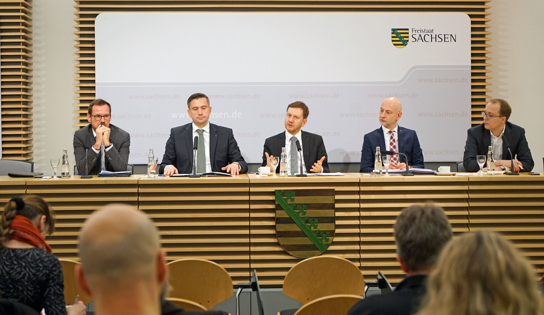 Männer sitzen auf einem Podest und halten eine Pressekonferenz.