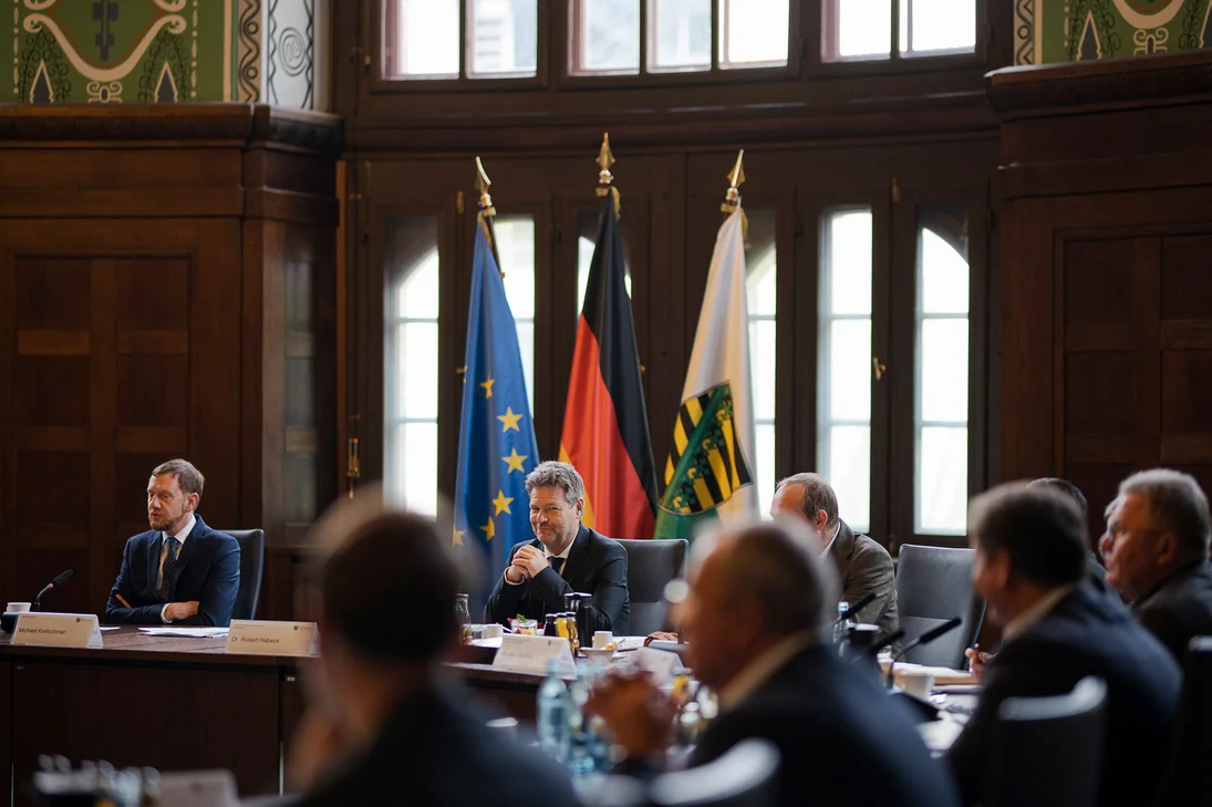 Männer sitzen an einem runden Tisch vor einer Fahnengalerie.
