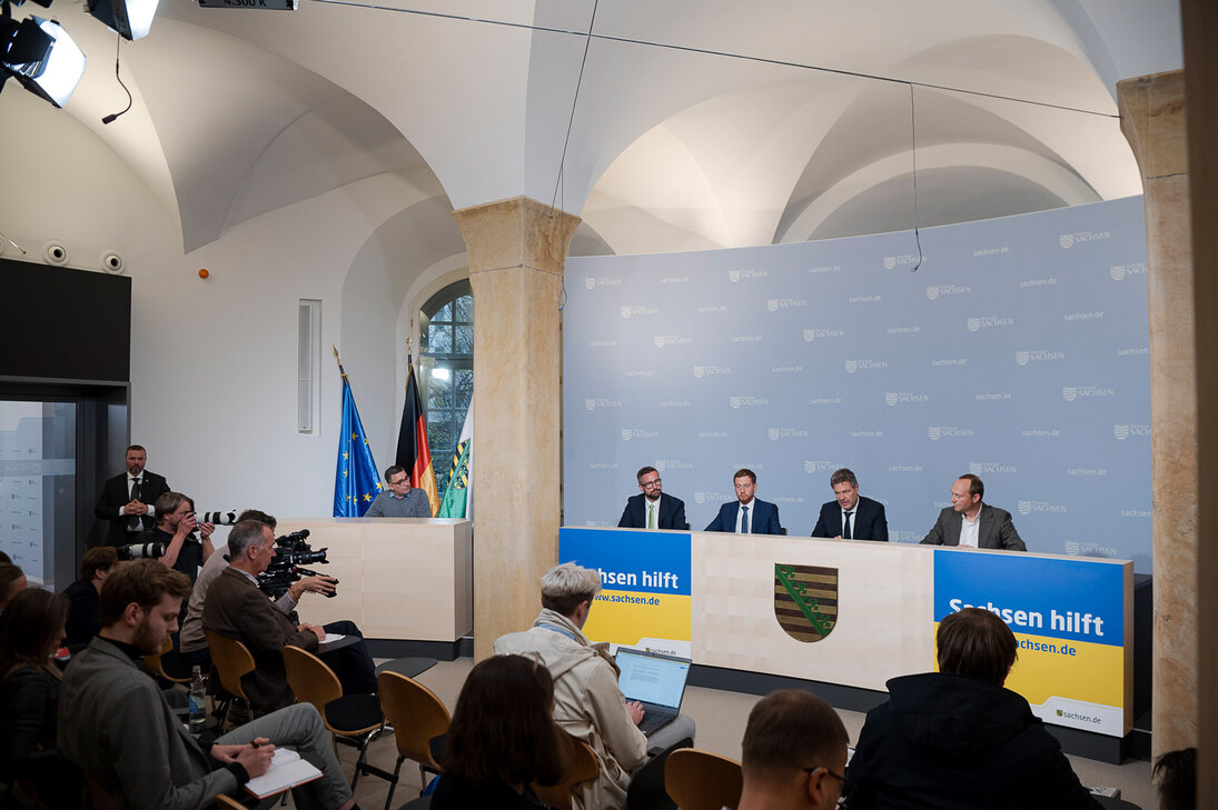 Vier Männer sitzen auf einem Podium. Vor stehen Menschen mit Kameras in der Hand.