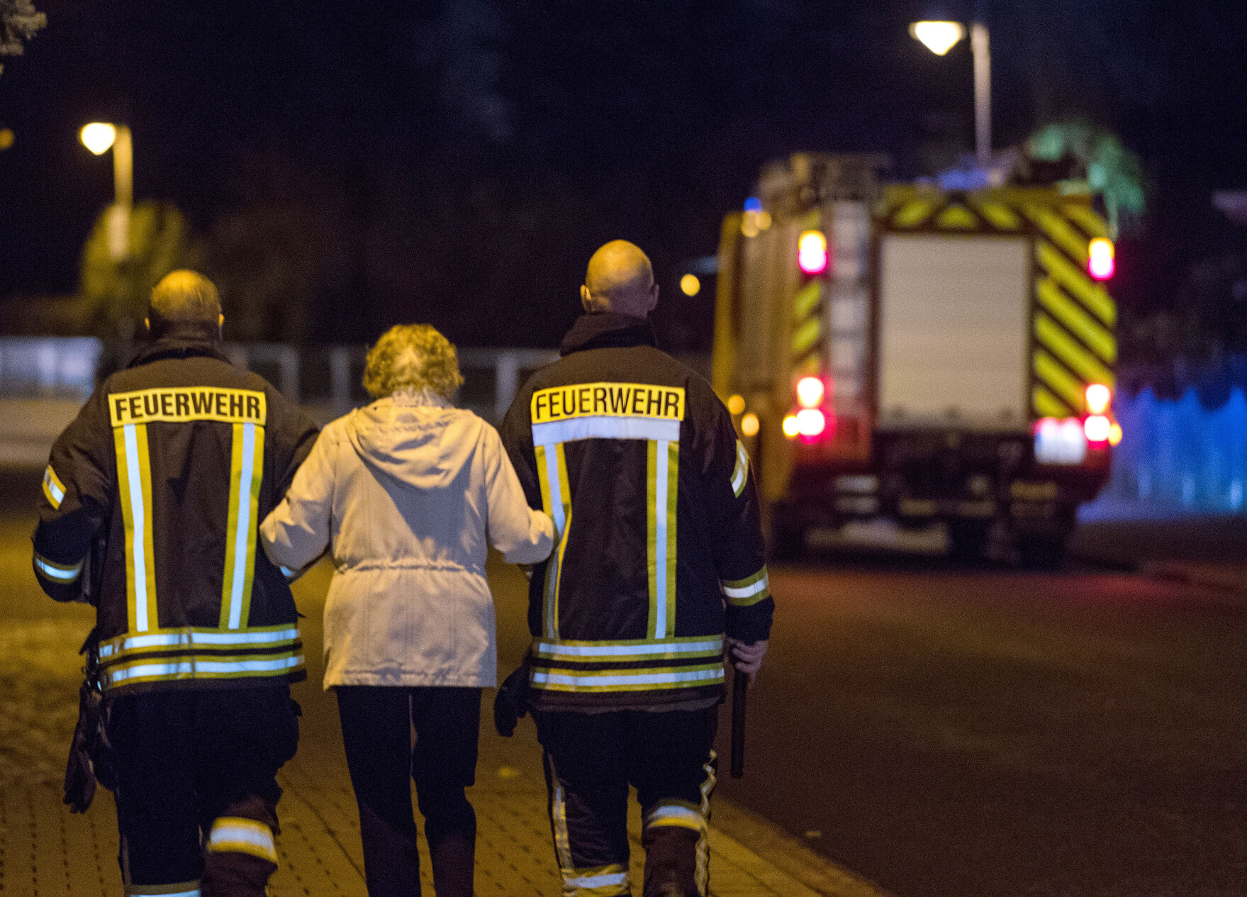 Zwei Feuerwehrmänner führen eine ältere Dame eine Straße entlang. Im Hintergrund sieht man unscharf ein Feuerwehrauto.