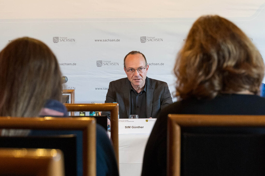 Ein Mann sitzt an einem Konferenztisch und spricht zu einer versammelten Personengruppe.