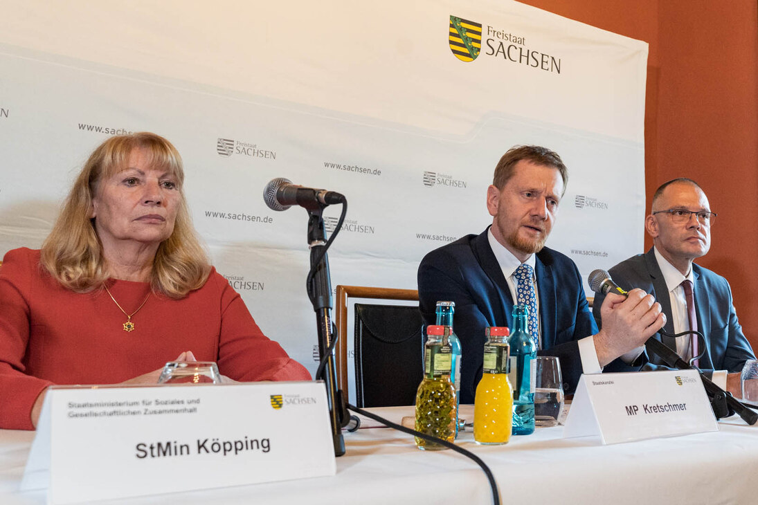 Zwei Männer und eine Frau sitzen an einem Tisch. Einer der Männer spricht in ein Mikrofon.