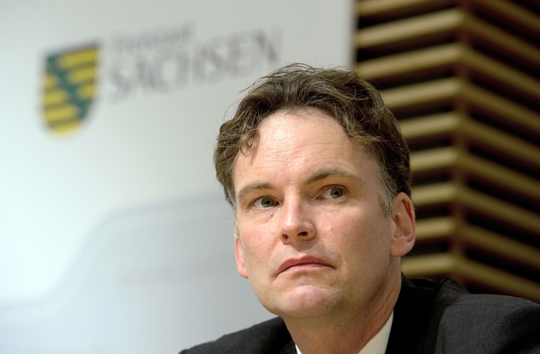 Ein Mann sitzt auf einem Podium vor den Logo des Freistaates Sachsen.