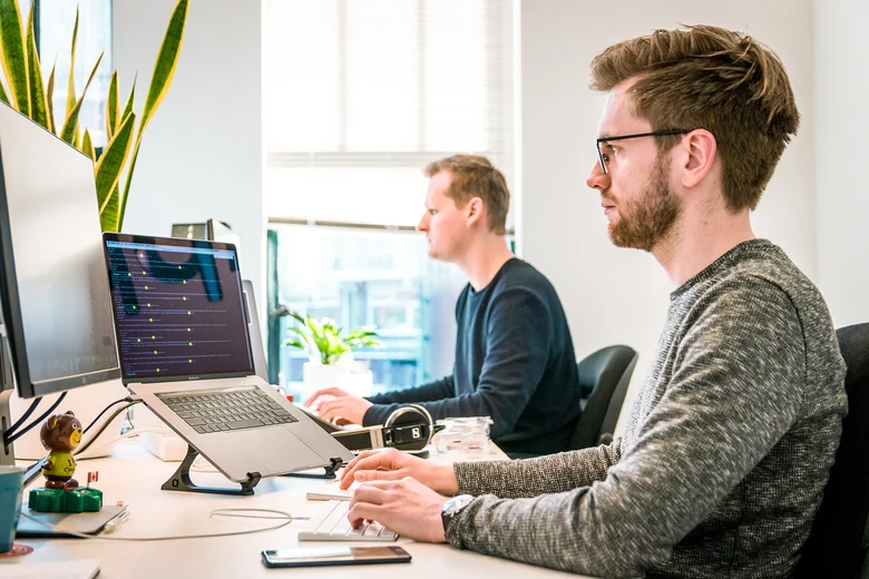 Zwei Männer sitzen an einem Schreibtisch und blicken auf Monitore.