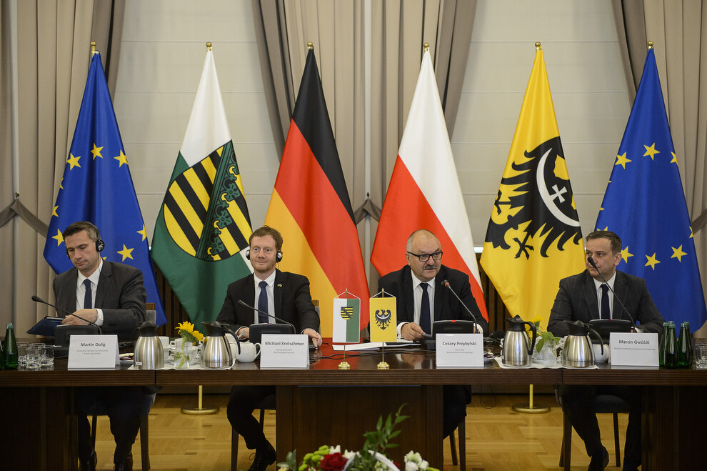 Männer sitzen an einem Tisch. Im Hintergrund steht eine Fahnengalerie.