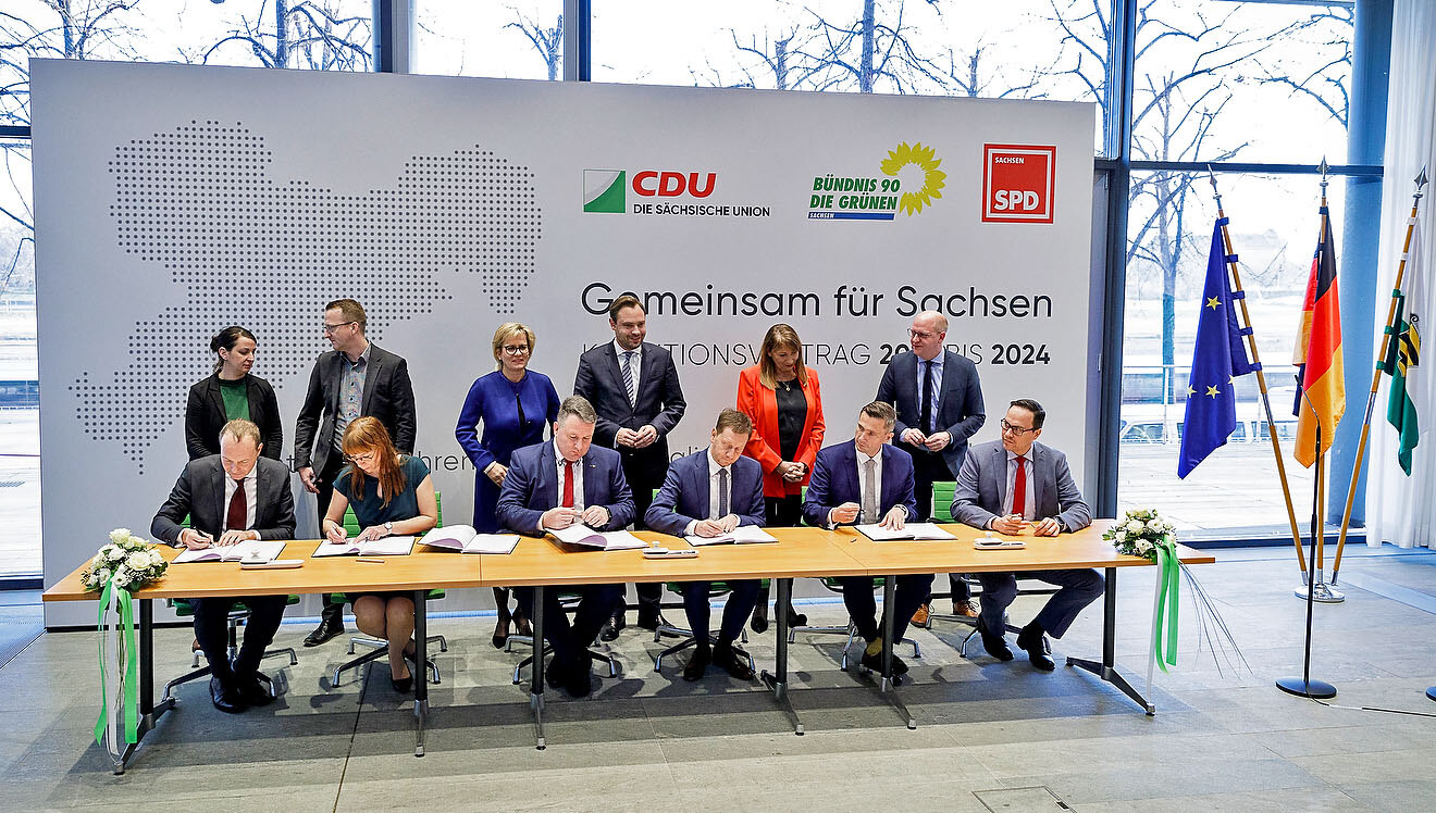 Männer und Frauen sitzen an einem Tisch und unterzeichnen einen Vertrag. Im Hintergrund steht eine Werbewand.